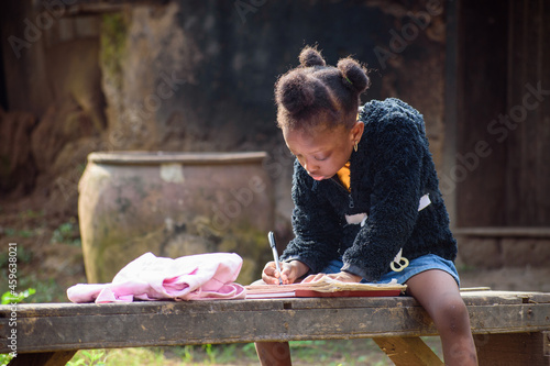 An African girl child or student sitting on a bench outside a village mud house, studying to attain excellence in her school, studies, career and education