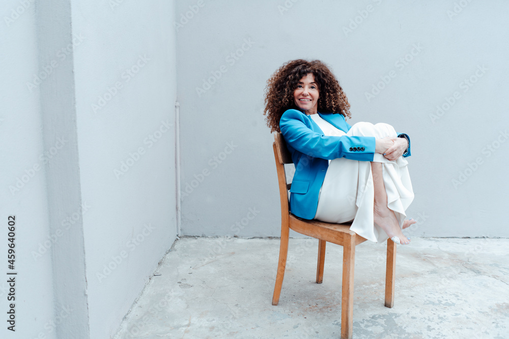 Businesswoman hugging knees while sitting on chair in front of gray wall Businesswoman hugging knees while sitting on chair in front of gray wall