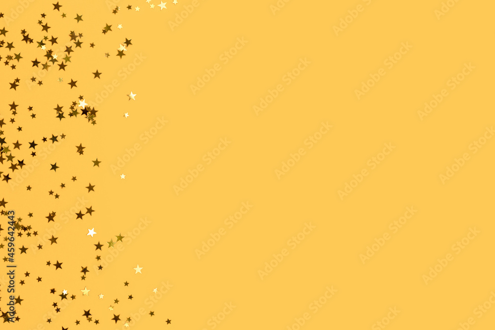 Glowing gold colored stars confetti scattered on a yellow background with copy space.