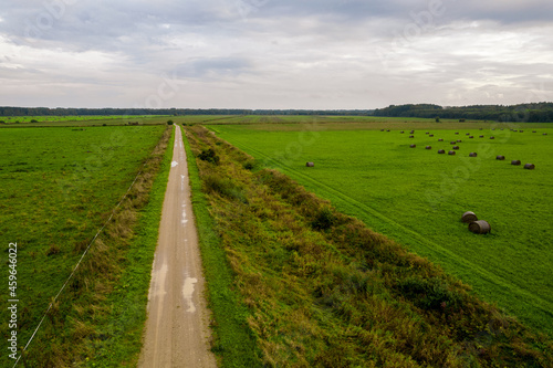 Aerial drone view shoot of rural dirty road leading through green farm lawn with straw bales and maize or rye plowed field and autumn forest. Agricultural parcels. Harvesting arable land  crop season