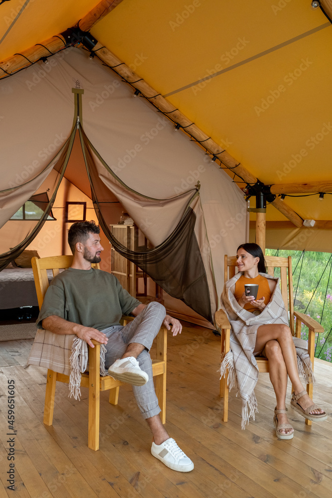 Restful young couple sitting in wooden armchairs on patio by glamping tent