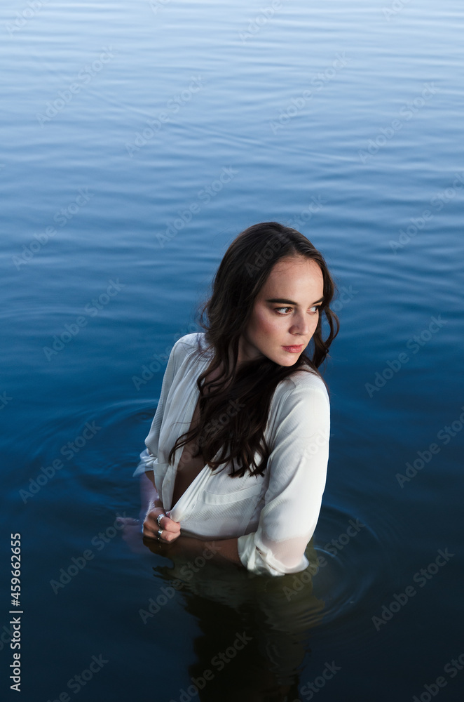 Thoughtful and calm young woman in white shirt is standing in water, looking away.