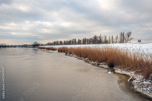 Fototapeta Naklejka Na Ścianę i Meble -  Snowy embankment along a frozen lake. Yellow reed plants are along the lake. The sky is covered with snow clouds. Two unrecognizable people are walking on the dike. It's winter in the Netherlands.