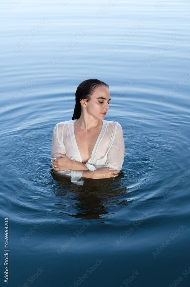 Fototapeta premium Young dreamy woman in white shirt standing in the lake, holding arms around herself, looking away.
