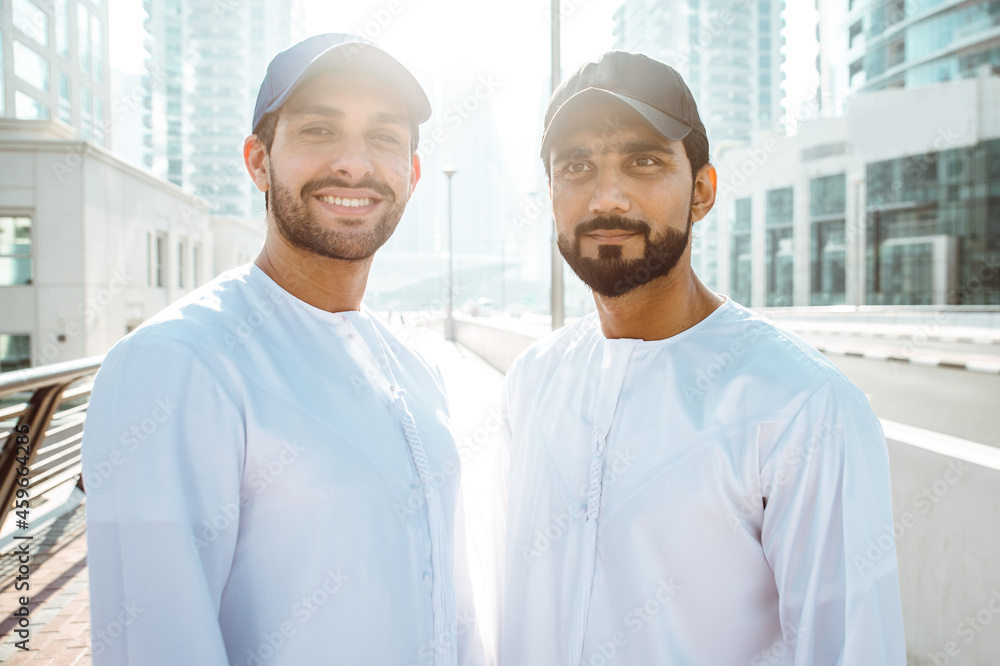 Two young men going out in Dubai. Friends wearing the kandura traditional male outfit and baseball hat in Marina Two young men going out in Dubai. Friends wearing the kandura traditional male outfit and baseball hat in Marina