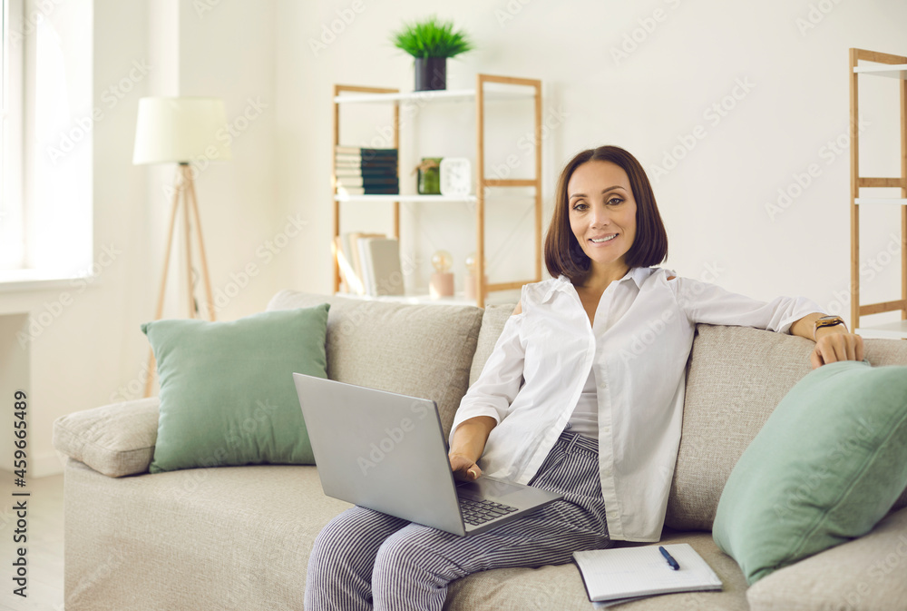 Portrait of senior stylish female freelancer working remotely using laptop and notepad. Happy beautiful caucasian woman sitting on sofa in her cozy comfortable home office. Remote business concept. Portrait of senior stylish female freelancer working remotely using laptop and notepad. Happy beautiful caucasian woman sitting on sofa in her cozy comfortable home office. Remote business concept.