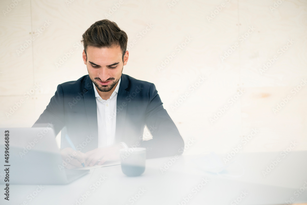 Young male professional writing while sitting at desk in office Young male professional writing while sitting at desk in office