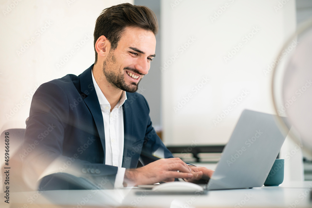 Happy young businessman using laptop while sitting at desk in office Happy young businessman using laptop while sitting at desk in office
