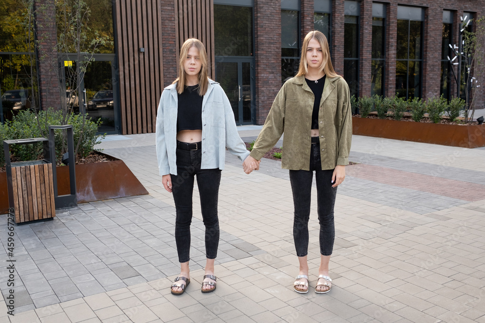 Blond twin girls in casualwear holding by hands while standing against modern building