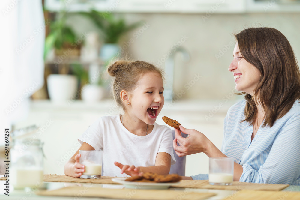 happy loving family, mom and daughter, playing sitting at table and having breakfast at home in morning. woman and girl eat oatmeal cookies and drink cow's milk, and have nice time together in kitchen happy loving family, mom and daughter, playing sitting at table and having breakfast at home in morning. woman and girl eat oatmeal cookies and drink cow's milk, and have nice time together in kitchen