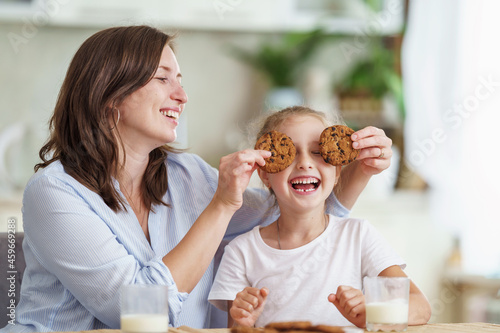 happy loving family  mom and daughter  playing sitting at table and having breakfast at home in morning. woman and girl eat oatmeal cookies and drink cow s milk  and have nice time together in kitchen