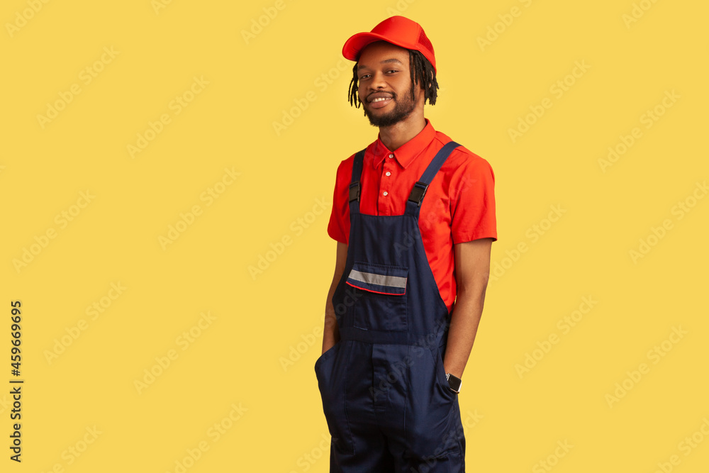 Confident satisfied handyman looking at camera, wearing uniform, being proud of done work, keeps hands in pockets, service industry. Indoor studio shot isolated on yellow background.
