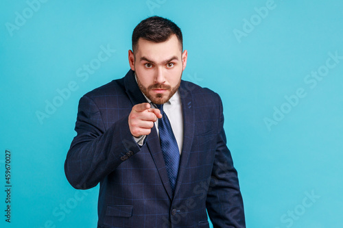 Serious bearded man wearing official style suit pointing finger, indicating direction to camera and looking annoyed, making choice. Indoor studio shot isolated on blue background.