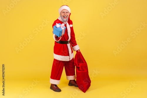 Full length portrait of elderly man with gray beard wearing santa claus costume standing with red bag with gifts and holding out Christmas present box. Indoor studio shot isolated on yellow background
