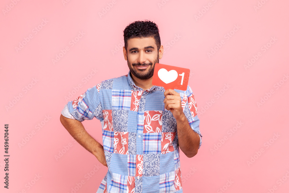 Like and follow interesting content! Portrait of smiling bearded man wearing blue shirt, holding social media heart icon, keeps hand on hip. Indoor studio shot isolated on pink background. Like and follow interesting content! Portrait of smiling bearded man wearing blue shirt, holding social media heart icon, keeps hand on hip. Indoor studio shot isolated on pink background.