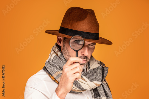 Male detective with magnifying glass, wearing fashionable scarf and brown hat, posing on yellow studio background.