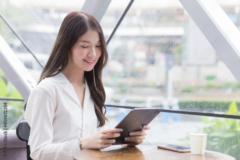 Young Asian professional business woman is looking at tablet or notepad in her hands while she is sitting at coffee shop among business background in the city.