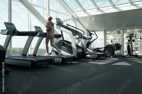 Horizontal long shot of modern Muslim woman wearing hijab exercising in gym running on treadmill, copy space