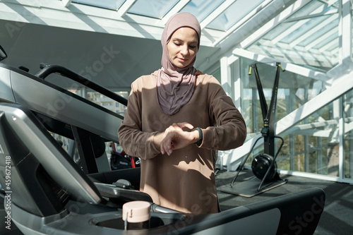 Horizontal medium portrait of modern young adult Muslim woman wearing hijab standing on treadmill in gym checking something on her activity tracker