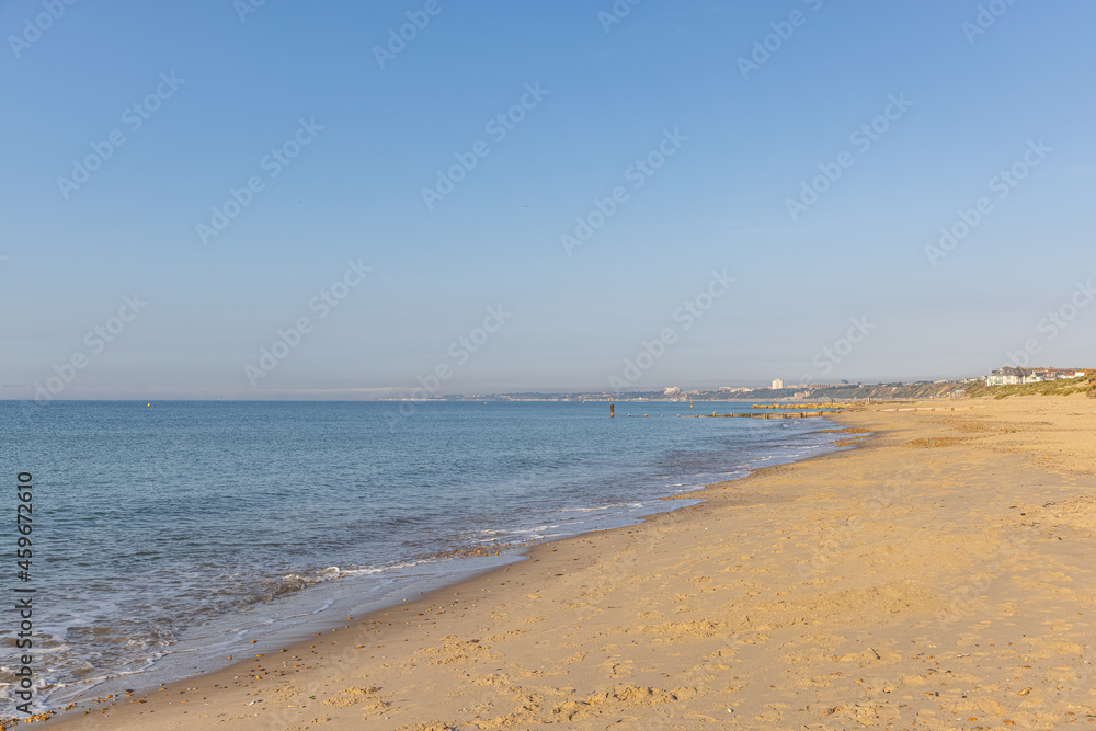 A scenic beautiful view of a sandy beach with blue sea, groynes, majestic cliffs, city buidldings and golden hours colors under a majestic blue sky and some white clouds