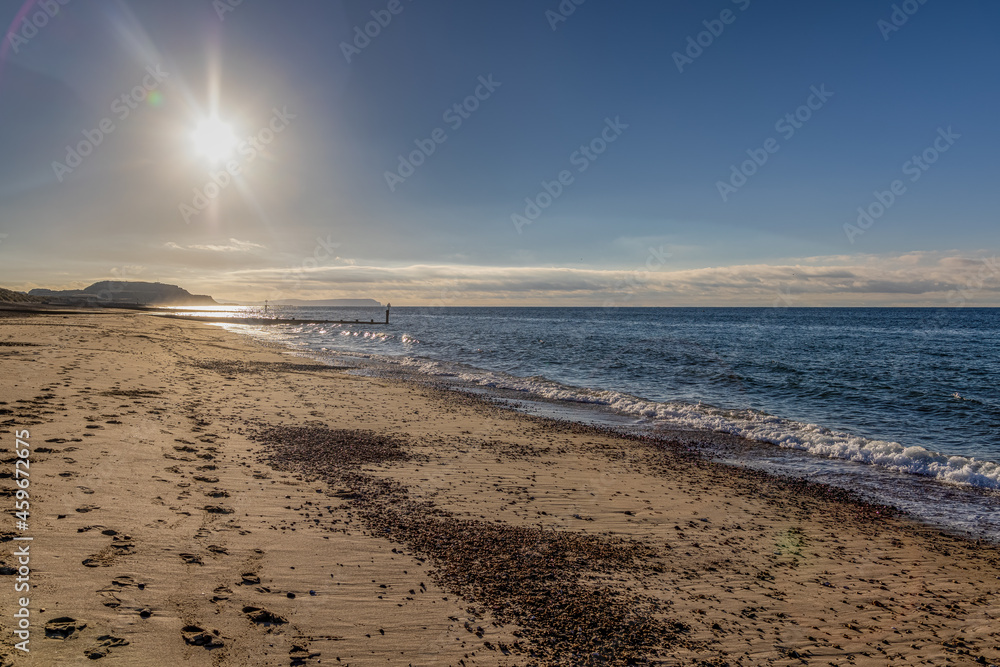 A scenic beautiful view of a sandy beach with beautiful blue sea, wooden groyne, majestic hill and shining sun under a beautiful blue sky and some white clouds