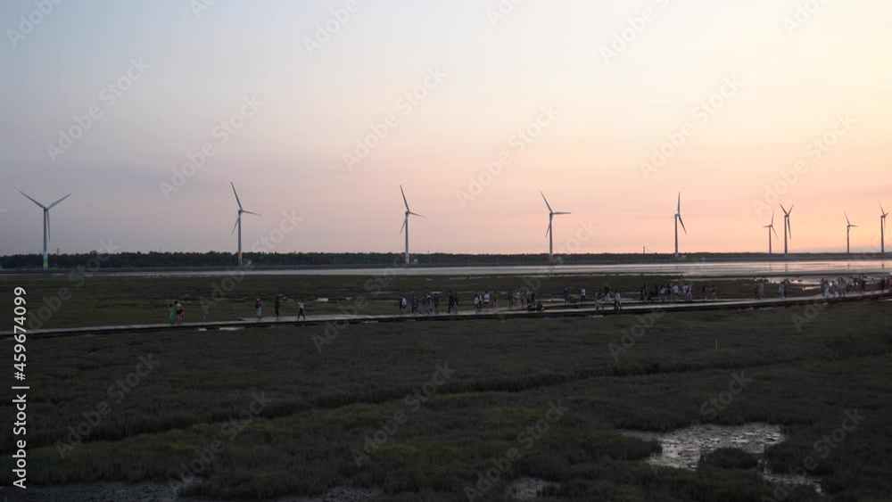 Tourists walking on the platform visiting well preserved remarkable gaomei wetland preservation area in Taichung, Taiwan, with a row of wind turbines in the background against diffused sunset color. Tourists walking on the platform visiting well preserved remarkable gaomei wetland preservation area in Taichung, Taiwan, with a row of wind turbines in the background against diffused sunset color.