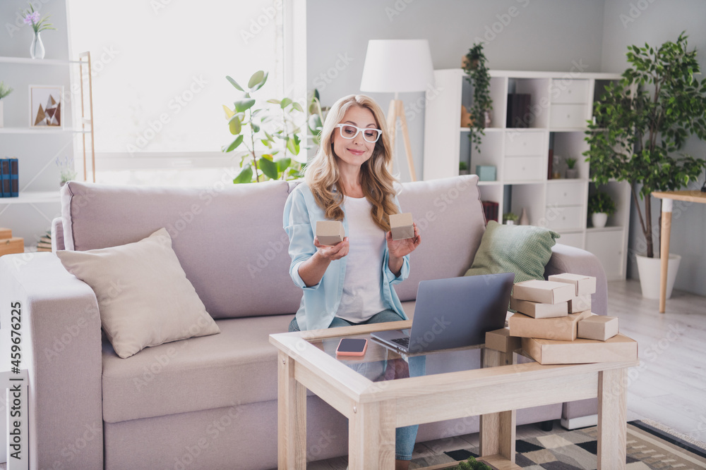 Photo of smiling businesswoman in glasses hold carton boxes working remote from house manager marketer use laptop Photo of smiling businesswoman in glasses hold carton boxes working remote from house manager marketer use laptop