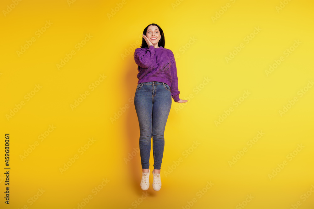 Full length view of attractive cheerful girl happy positive smile excited amazed hand touch cheek jump isolated over yellow color background Full length view of attractive cheerful girl happy positive smile excited amazed hand touch cheek jump isolated over yellow color background
