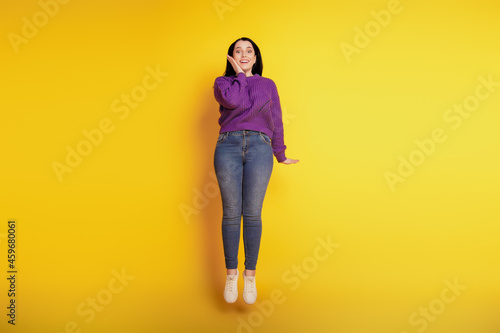 Full length view of attractive cheerful girl happy positive smile excited amazed hand touch cheek jump isolated over yellow color background