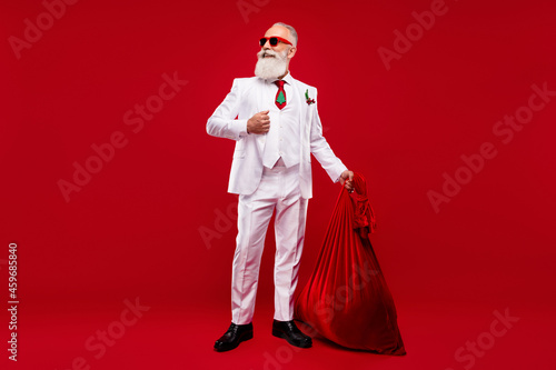 Full length photo of happy dreamy old man look empty space hold bag santa claus isolated on red color background