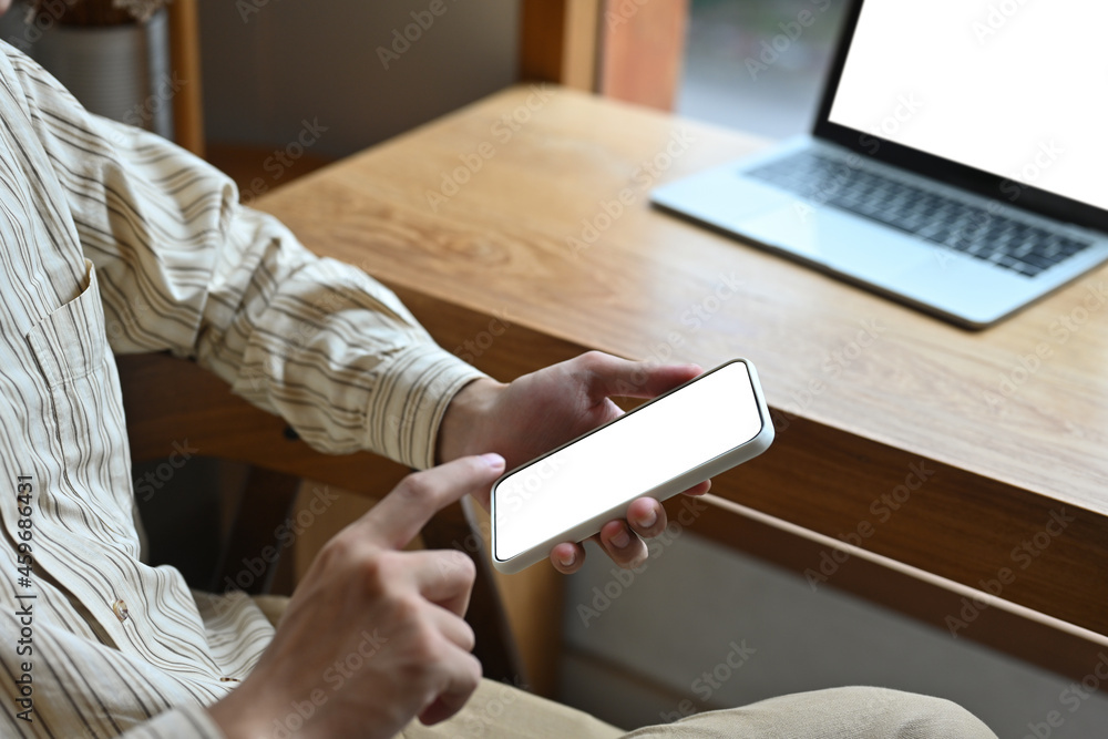 Cropped image of a man's hand  holding a white screen smartphone with a computer laptop place on the wooden working desk as a background.