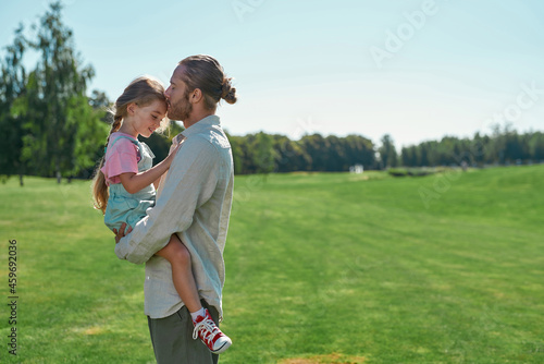 Caring young dad holding his little daughter and gently kissing her while spending time together in the park on a summer day