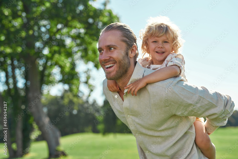 Happy father playing in the park, smiling together with his little son sitting on his back Happy father playing in the park, smiling together with his little son sitting on his back