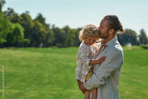 Caring young dad holding his little son and gently kissing him while spending time together in the park on a summer day
