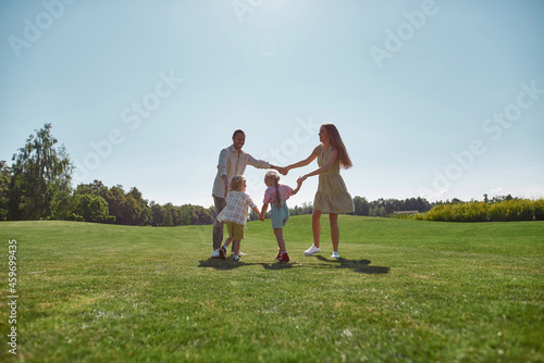 Active parents spending time together with two little kids, boy and girl in green park on a summer day. Happy family enjoying leisure activity