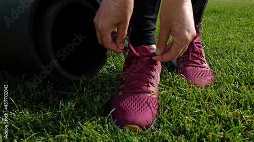 Young woman ties the laces on the pink sneaker. Woman preparing for yoga asana or fitness workout on green lawn in the backyard or park. Warm up before running. Healthy lifestyle. 