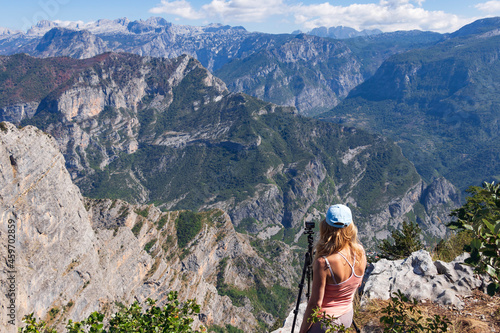 A young woman traveler stands on top of a mountain looking down at the Grlo Sokolovo Gorge and the surrounding mountains, next to an action camera on a tripod, capturing a breathtaking time-lapse view