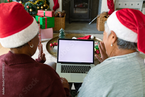 Two diverse senior female friends wearing santa hats using laptop with copy space at christmas time