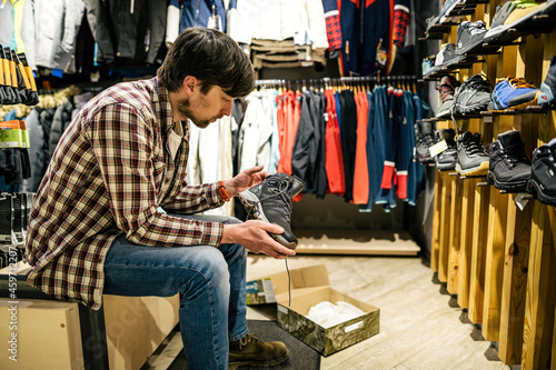 Man shopping in an outdoor store. Male buying mountain boots. Shopper in sports store select hiking shoes. Customer purchase of footwear for adventures. Man trying mountaineering boots at travel shop