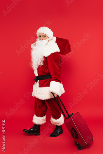 Santa claus in costume holding suitcase and sack on red background