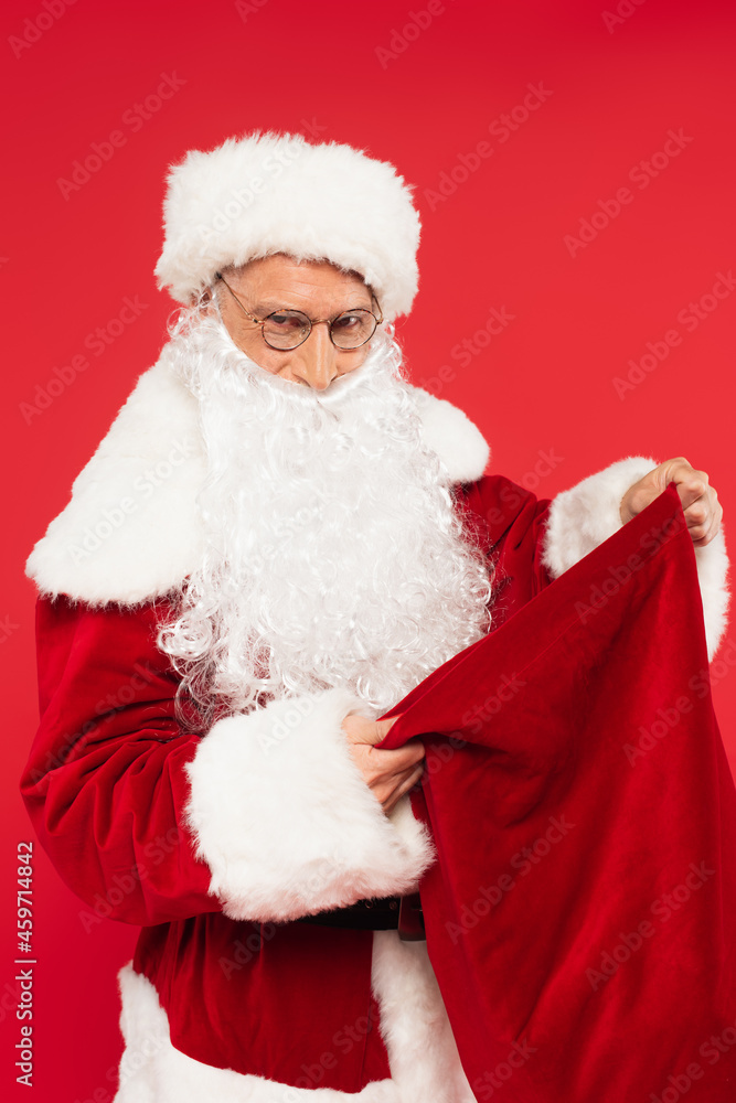 Santa in eyeglasses looking at camera and holding sack isolated on red Santa in eyeglasses looking at camera and holding sack isolated on red