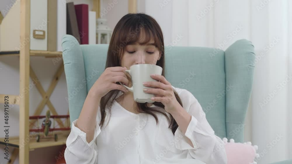 portrait beautiful asian woman is looking at the camera with a smile while sipping hot tea on an armchair at home. portrait beautiful asian woman is looking at the camera with a smile while sipping hot tea on an armchair at home.