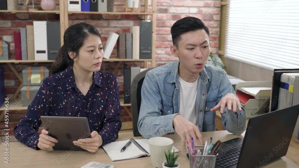 asian man is pointing at the tablet in his coworker’s hand while expressing thoughts by consulting his laptop during a brainstorm meeting in office.
