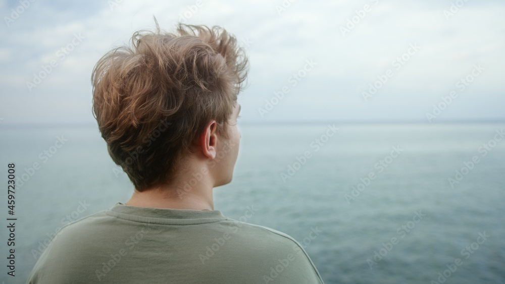 Young handsome blond boy puts his hands behind his neck and looks towards the horizon, deep in thought
