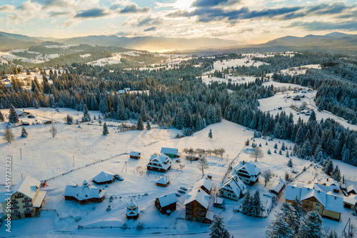 Aerial winter landscape with small village houses between snow covered forest in cold mountains in the evening.