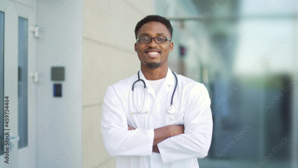 Close up portrait African American doctor looking at camera with arms crossed background of modern hospital outside. Confident happy medic outdoors. male scientist therapist wearing scrubs near clinic Close up portrait African American doctor looking at camera with arms crossed background of modern hospital outside. Confident happy medic outdoors. male scientist therapist wearing scrubs near clinic