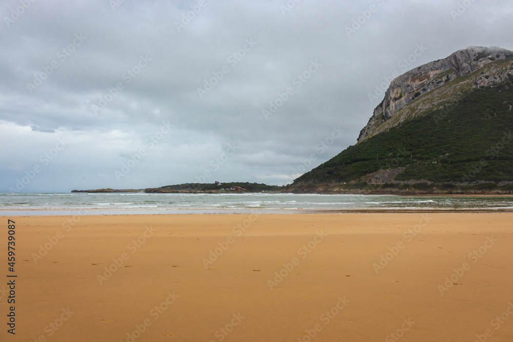 Wide surf beach with mountain in clouds. Autumn landscape with empty coastline. Turquoise seascape. Camino de Santiago landscape. Tourism in Spain. Travel inspiration concept. Beauty in coastal nature Wide surf beach with mountain in clouds. Autumn landscape with empty coastline. Turquoise seascape. Camino de Santiago landscape. Tourism in Spain. Travel inspiration concept. Beauty in coastal nature