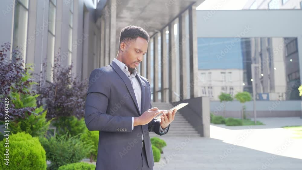 Young african american businessman in formal business suit standing working with tablet in hands on background modern office building outside. Man using smartphone or mobile phone outdoors city street Young african american businessman in formal business suit standing working with tablet in hands on background modern office building outside. Man using smartphone or mobile phone outdoors city street