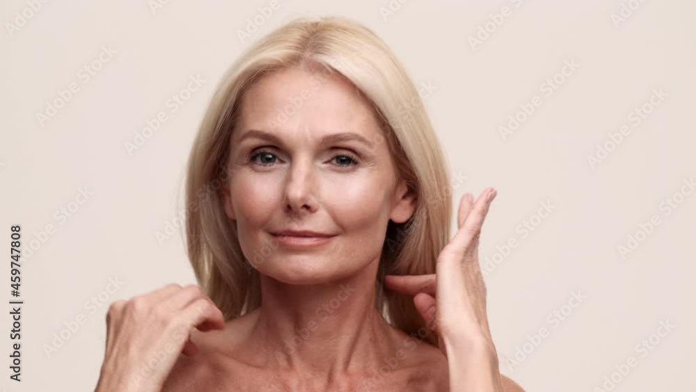A close-up view of the positive mature beautiful woman correcting her hairstyle at the camera in the light beige studio A close-up view of the positive mature beautiful woman correcting her hairstyle at the camera in the light beige studio