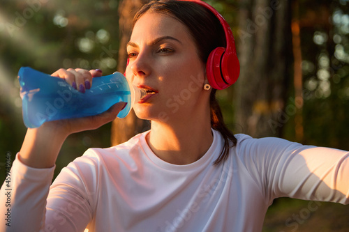 Close-up of a beautiful female fitness athlete in red wireless headphones who drinks water from a reusable soft sports bottle after a workout.